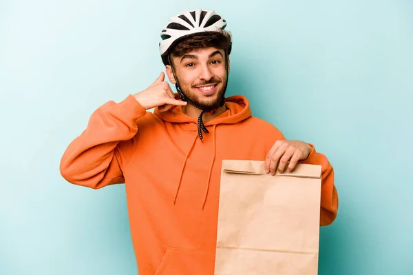 Young hispanic man wearing a helmet bike holding a take away food isolated on blue background showing a mobile phone call gesture with fingers.