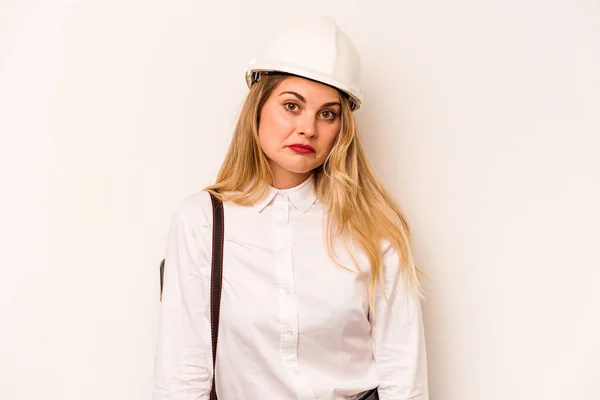 Young architect woman with helmet and holding blueprints isolated on white background shrugs shoulders and open eyes confused.