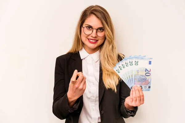 Young business caucasian woman holding a banknotes isolated on white background pointing with finger at you as if inviting come closer.