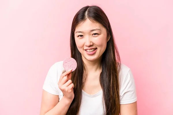 年轻的亚洲女人 手里拿着用粉色背景笑着隔离起来的面部海绵 玩得很开心 — 图库照片