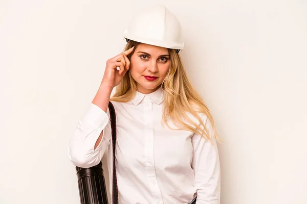 Young architect woman with helmet and holding blueprints isolated on white background pointing temple with finger, thinking, focused on a task.