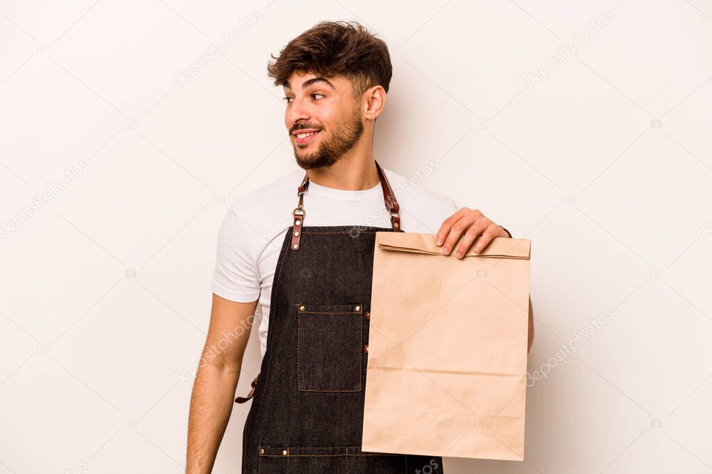 Young hispanic clerk man holding a take away bag isolated on white background looks aside smiling, cheerful and pleasant.
