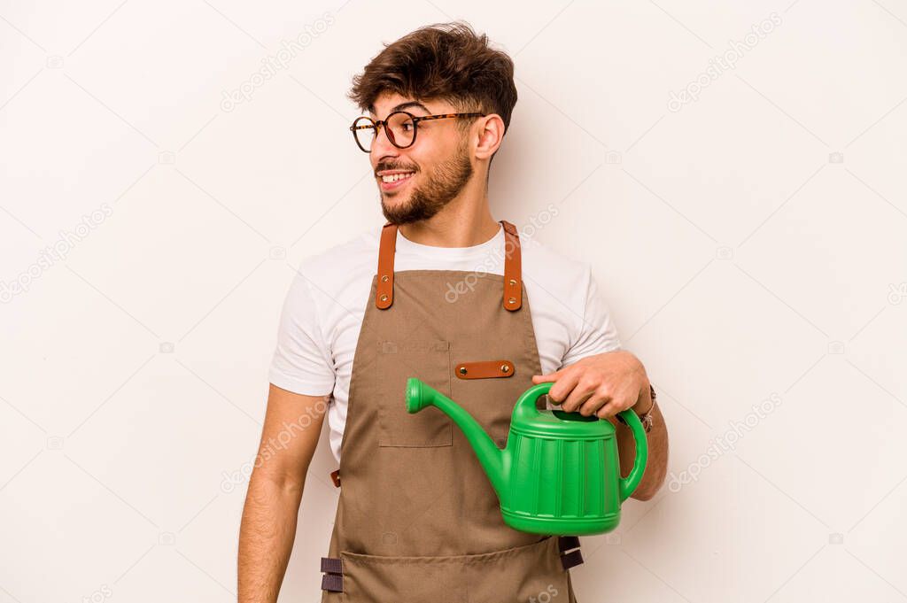 Young gardener hispanic man holding a watering can isolated on white background looks aside smiling, cheerful and pleasant.