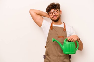 Genç bahçıvan, elinde su dolu bir teneke tutarak beyaz arka planda izole edilmiş kafasının arkasına dokunuyor, düşünüyor ve bir seçim yapıyor..