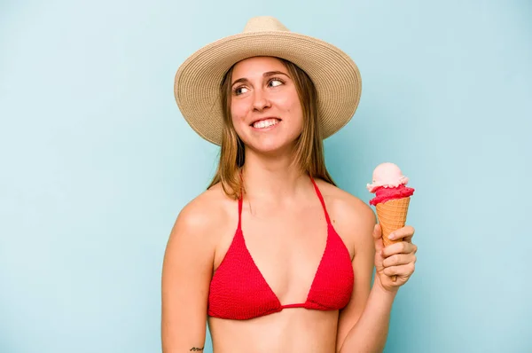 身穿比基尼 拿着蓝色背景的冰淇淋的年轻的高加索女人 看上去面带微笑 快乐而愉快 — 图库照片