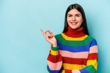 Mavi arka planda izole edilmiş genç beyaz kadın işaret parmağıyla işaret ederek gülümsüyor..
