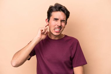 Beyazlı genç bir adam bej arka planda izole edilmiş parmaklarıyla kulaklarını kapatıyor, gürültülü bir ortam yüzünden stresli ve çaresiz..