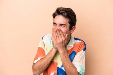 Beyazlı genç bir adam bej arka planda izole edilmiş bir şekilde bir şeye gülüyor, ağzı ellerle örtüyor..