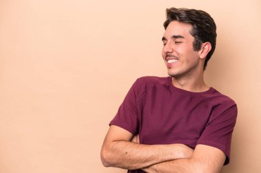 Beyaz tenli genç adam bej arka planda izole edilmiş. Kollarını kavuşturup gülümsüyor..