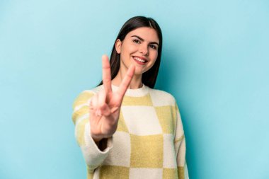 Mavi arka planda izole edilmiş genç beyaz kadın neşeli ve kaygısız parmakları olan bir barış sembolü gösteriyor..