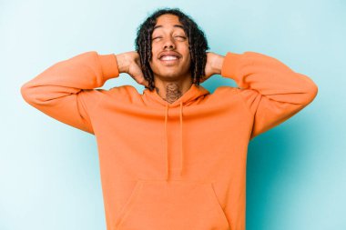 Genç Afrikalı Amerikalı bir adam mavi arka planda izole edilmiş, kendinden emin, elleri başının arkasında..