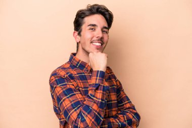 Beyaz tenli genç adam bej arka planda izole edilmiş mutlu ve kendinden emin gülümsüyor, elleriyle çenesine dokunuyor..