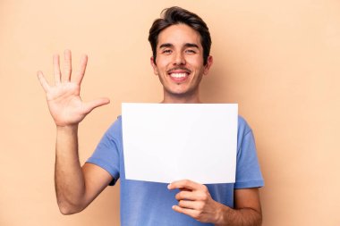 Beyazlı genç bir adam bej arka planda izole edilmiş bir pankart tutuyor. Beş numarayı parmaklarıyla gülümsüyor..