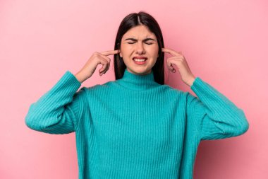 Pembe arka planda izole edilmiş genç beyaz bir kadın parmaklarıyla kulaklarını kapatıyor, gürültülü bir ortam yüzünden stresli ve çaresiz..
