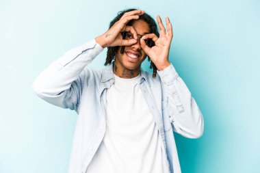 Genç Afro-Amerikalı adam mavi arka planda izole edilmiş, gözlerinde sorun yok işareti var.