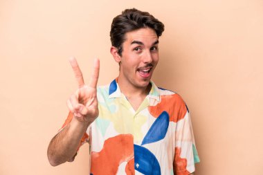 Beyazlı genç bir adam bej arka planda izole edilmiş. İki numarayı parmaklarıyla gösteriyor..