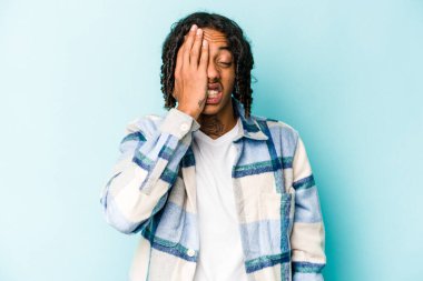 Genç Afrikalı Amerikalı adam mavi arka planda izole edilmiş. Yüzünün yarısını avuç içiyle kaplayarak eğleniyor..