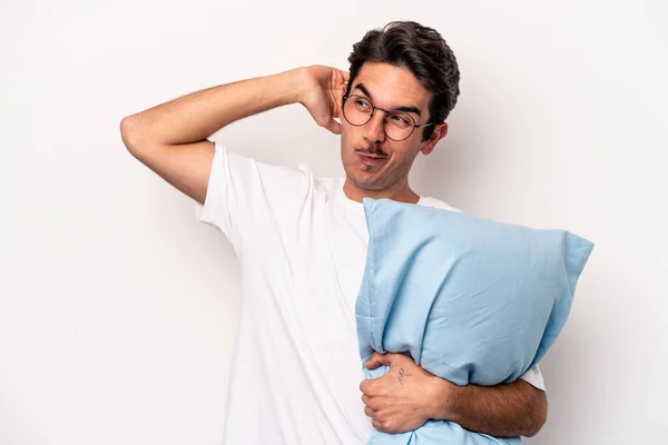 頭の後ろに触れる白い背景に隔離された枕を保持パジャマを身に着けている若い白人男性 思考と選択をする — ストック写真