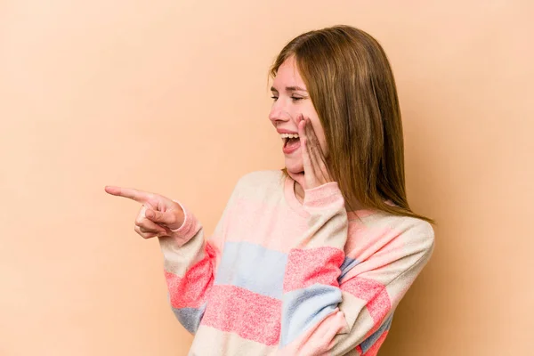 Young English woman isolated on beige background saying a gossip, pointing to side reporting something.