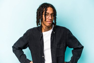 Mavi arka planda izole edilmiş, hoşnutsuzca kaşlarını çatmış, kollarını kavuşturmuş genç Afrikalı Amerikalı bir adam..