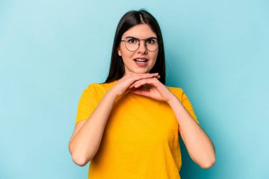 Kafkasyalı genç bir kadın mavi arka planda izole edilmiş şans için dua ediyor, şaşırmış ve ağzını açıp önüne bakıyor..