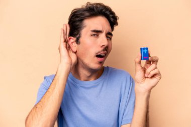 Beyazlı genç bir adam bej arka planda izole edilmiş bir bateriyi tutuyor ve bir dedikoduyu dinlemeye çalışıyor..