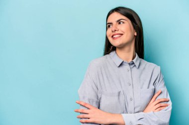 Mavi arka planda izole edilmiş genç beyaz kadın kollarını kavuşturarak gülümsüyor..