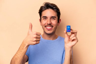Beyazlı genç bir adam bej arka planda izole edilmiş bir bateriyi tutuyor gülümsüyor ve başparmağını kaldırıyor.