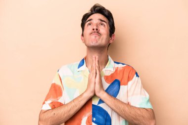 Beyaz tenli genç, bej arka planda izole edilmiş, el ele tutuşmuş, ağzı yakınında dua eden, kendinden emin..