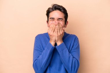 Beyazlı genç bir adam bej arka planda izole edilmiş bir şekilde bir şeye gülüyor, ağzı ellerle örtüyor..