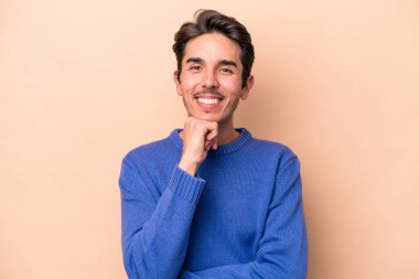 Beyaz tenli genç adam bej arka planda izole edilmiş mutlu ve kendinden emin gülümsüyor, elleriyle çenesine dokunuyor..