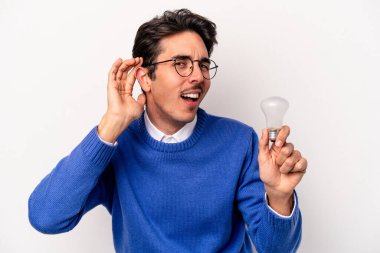 Beyaz arka planda izole edilmiş bir ampul tutan genç beyaz bir adam bir dedikoduyu dinlemeye çalışıyor..