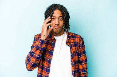 Genç Afrikalı Amerikalı bir adam mavi arka planda izole edilmiş ve parmakları dudaklarında bir sır saklıyor..