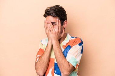 Beyazlı genç bir adam bej arka planda izole edilmiş. Kameraya parmaklarıyla göz kırpıyor, yüzünü kapatmaktan utanıyor..