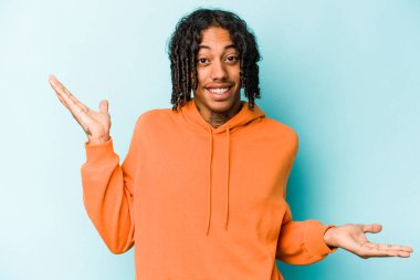 Genç Afrikalı Amerikalı bir adam mavi arka planda izole edilmiş. Kafası karışmış ve omuz silkiyor..