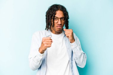 Mavi arka planda izole edilmiş genç Afro-Amerikalı adam yumruğunu kameraya gösteriyor, agresif yüz ifadesi..