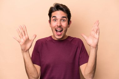 Beyaz tenli genç adam bej arka planda izole edilmiş. Hoş bir sürpriz bekliyor. Heyecanlı ve el kaldırıyor..