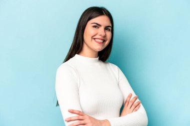 Mavi arka planda izole edilmiş genç beyaz kadın kendini güvende hissediyor, kararlılıkla kollarını kavuşturuyor..