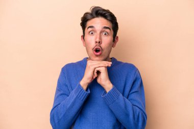 Beyaz tenli genç adam bej arka planda izole edilmiş şans için dua ediyor, şaşırmış ve ağzını açıp önüne bakıyor..