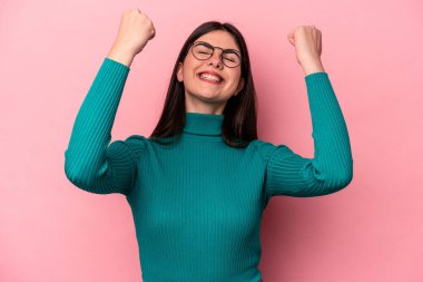 Pembe arka planda izole edilmiş genç beyaz kadın zaferi, tutkuyu ve coşkuyu kutluyor, mutlu bir ifade..