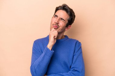 Beyaz tenli genç adam bej arka planda izole edilmiş düşünüyor ve yukarı bakıyor, yansıtıyor, düşünüyor, bir fantezi kuruyor..