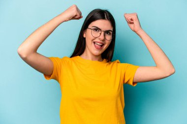 Mavi arka planda izole edilmiş genç beyaz kadın güçlü bir hareket sergiliyor kollarıyla, kadınsı gücün sembolü.