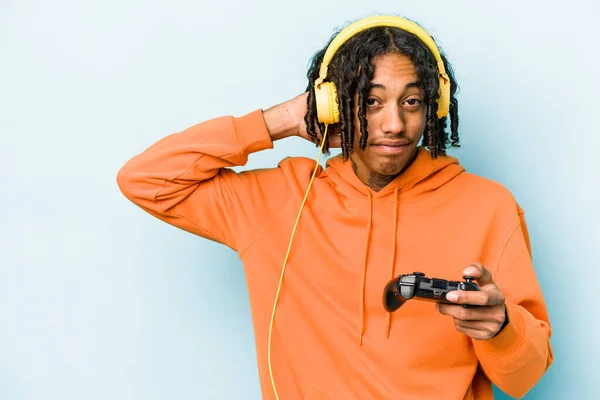 Young African American man playing with a video game controller isolated on blue background touching back of head, thinking and making a choice.