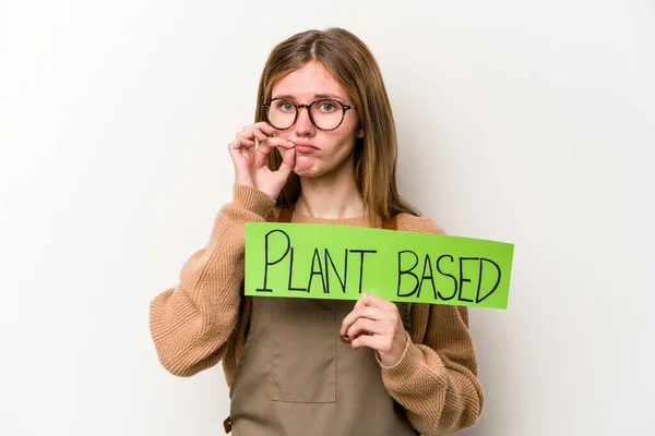 Young gardener woman holding a plan based placard isolated on white background with fingers on lips keeping a secret.