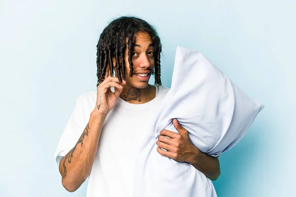 Young African American man holding a pillow isolated on blue background trying to listening a gossip.