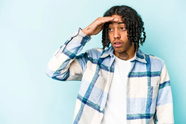 Genç Afrikalı Amerikalı adam mavi arka planda izole edilmiş, uzaklara bakıyor ve elini alnına koyuyor..