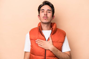 Beyaz tenli genç adam bej arka planda izole edilmiş yemin ediyor, elini göğsüne koyuyor..