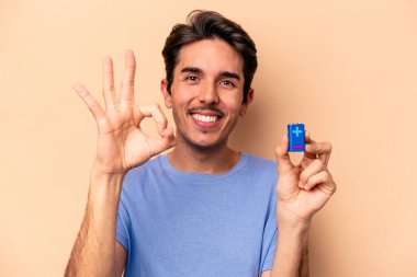 Beyazlı genç bir adam bej arka planda izole edilmiş bir bateriyi tutuyor neşeli ve kendinden emin bir şekilde iyi bir hareket gösteriyor..