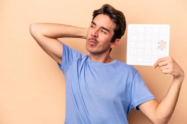 Kafkasyalı genç bir adam bej arka planda izole edilmiş bir yapboz tutuyor kafasının arkasına dokunuyor, düşünüyor ve bir seçim yapıyor..