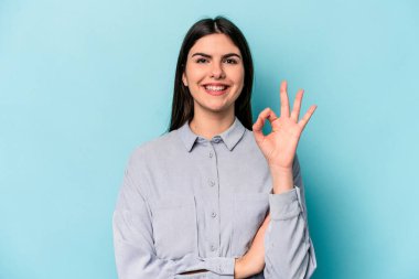 Kafkasyalı genç bir kadın mavi arka planda izole edilmiş bir şekilde bir göz kırpıyor ve elleriyle iyi bir jest yapıyor..